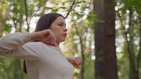 woman warms up before cardio workout in forest. lady does arms turning exercise preparing for long run surrounded by dense trees at sunlight. training in nature at sunset