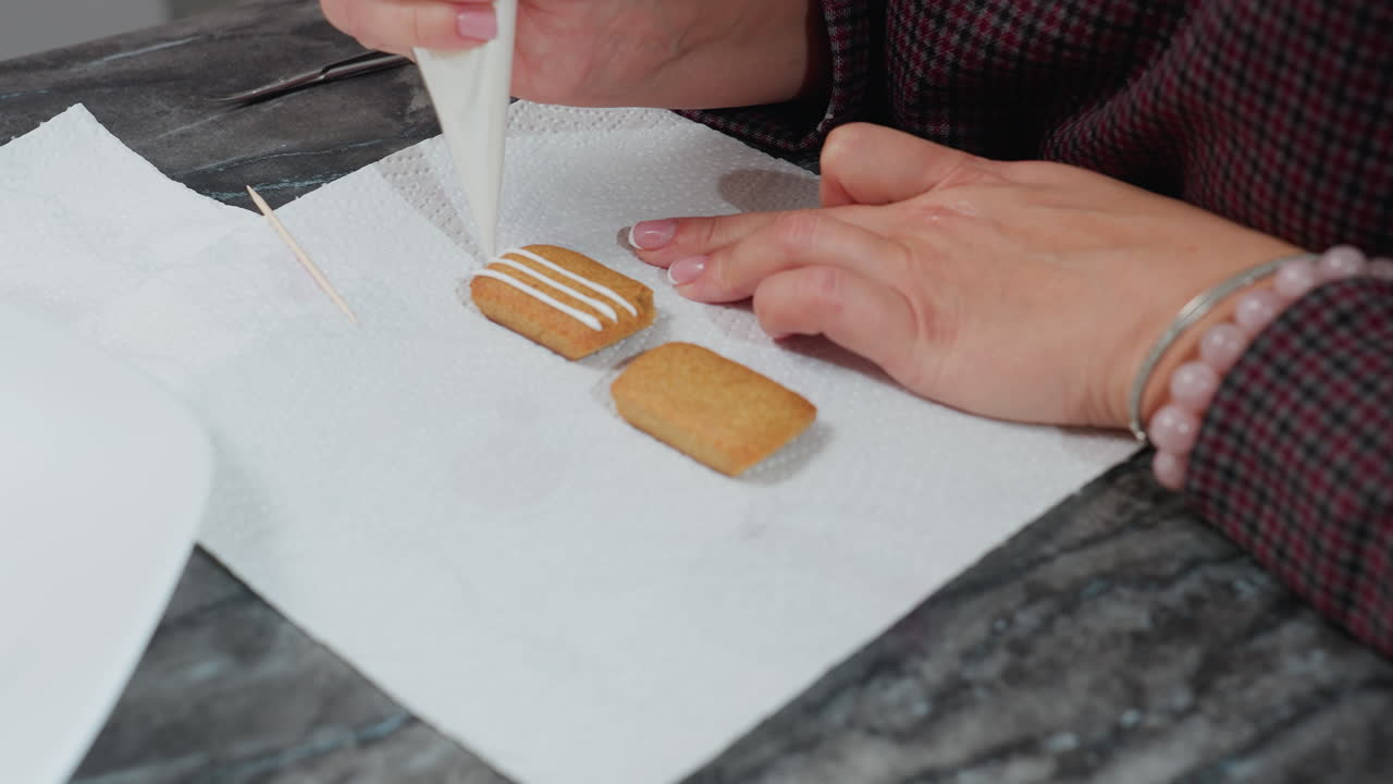 primer plano de una panadería decorando una galleta con glaseado blanco en papel de pañuelo, mano adornada con una pulsera de cuentas, palillo de dientes cerca para detalles precisos