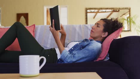 Mujer-Caucásica-Leyendo-Un-Libro,-Tumbada-En-El-Sofá-De-Casa