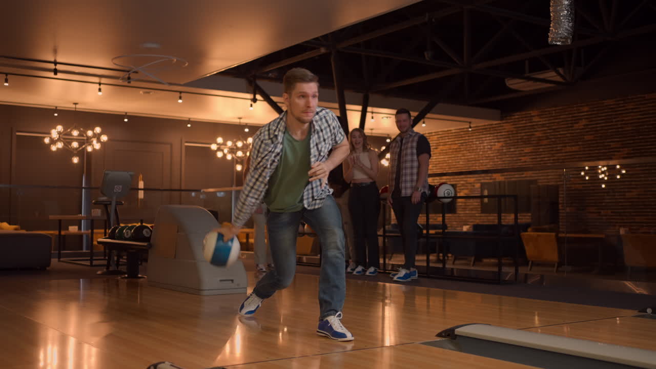 A man in a bowling alley throws a ball on the track and knocks out a shot in slow motion and jumps and dances for joy. A group of multi-ethnic friends play bowling together clapping happy supporting
