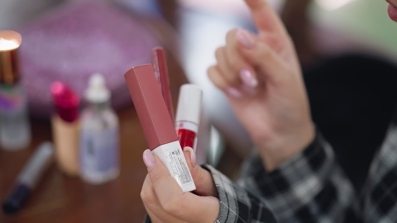 частичный вид женщины, держащей и исследующей несколько предметов макияжа в руке, фон включает в себя размытые косметические продукты на деревянном столе, фиксируя фокус на выборе косметики