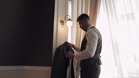 Handsome-groom-businessman-taking-and-preparing-to-wear-gray-jacket-at-home-in-living-room