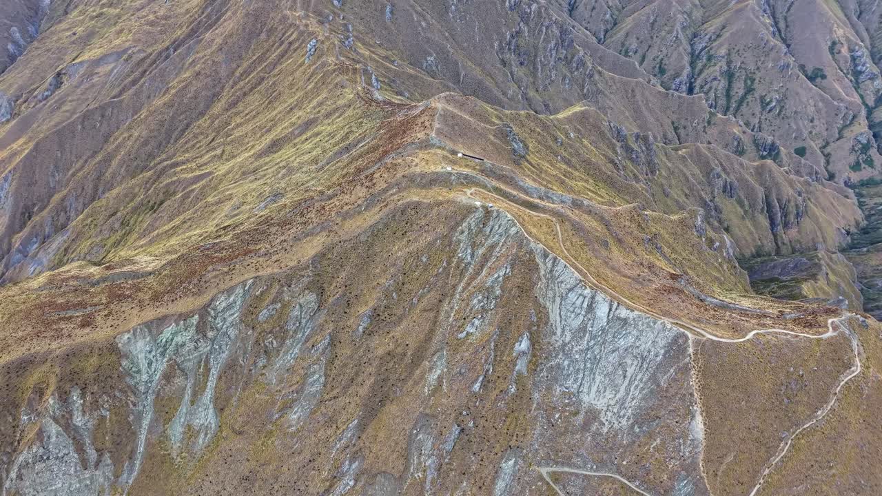 Drone in a high top-down angle flying toward the summit of Roys Peak, showing the winding hiking trail and rocky alpine terrain