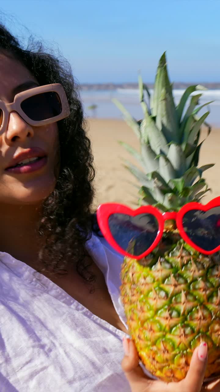 Mulher feliz posando com abacaxi e usando óculos de sol de coração na praia.
