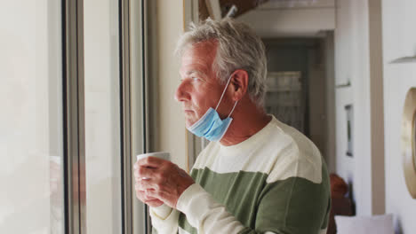 Älterer-Kaukasischer-Mann-Mit-Abgesenkter-Gesichtsmaske-Trinkt-Kaffee,-Während-Er-Zu-Hause-Aus-Dem-Fenster-Schaut