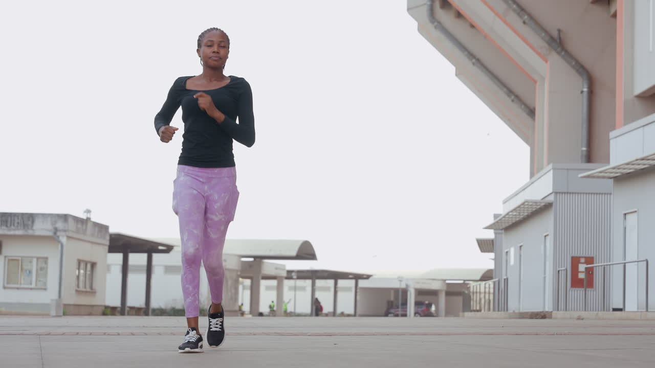 Mulher correndo em direção à câmera, com close-up da ação das pernas em leggings roxas e tênis. Forma atlética e respiração visíveis durante um sprint energético perto de um armazém, uma atleta confiante testando sua velocidade.