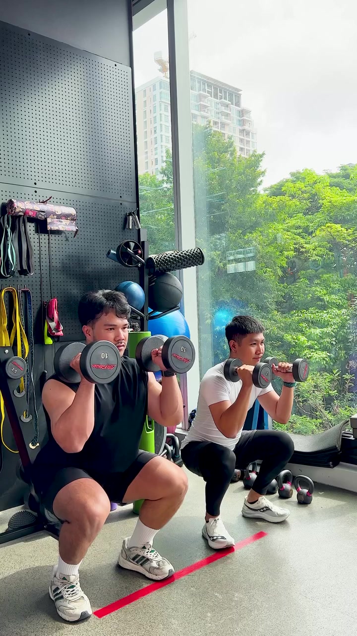 Dua pria melakukan squat dumbbell secara sinkron di gym yang terang dengan jendela kota yang besar