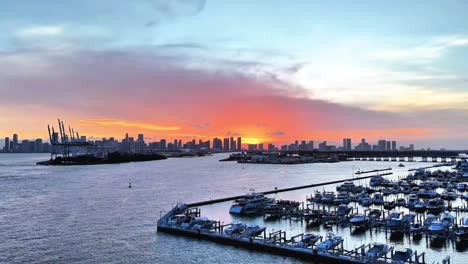 drone ascends towards the miami downtown skyline with a setting sun behind it, passing a marina full of boats and the port of miami