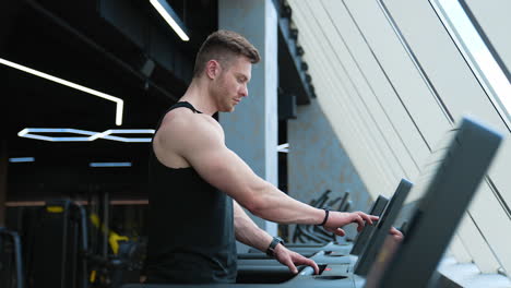 sportsman in fitness club. young handsome athlete switches on electronic treadmill in the gym. muscular man exercising on a simulator. profile view.