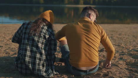 young female and male friends sharing moments sitting on the beach seen from behind.