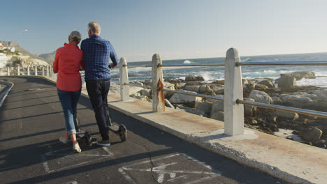 Senior-couple-walking-next-to-electronic-scooter-alongside-beach