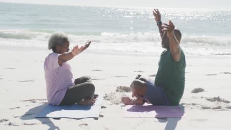 happy senior african american couple doing yoga and meditating at beach, in slow motion