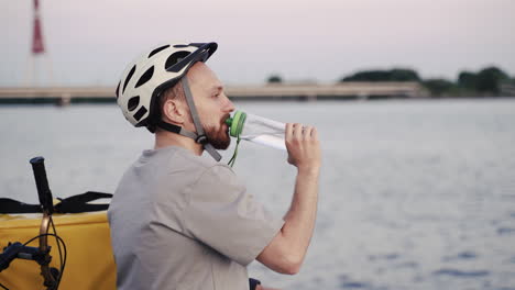 el repartidor de comida agotado sentado al lado de un río al atardecer bebe un poco de agua de una botella de plástico durante su descanso