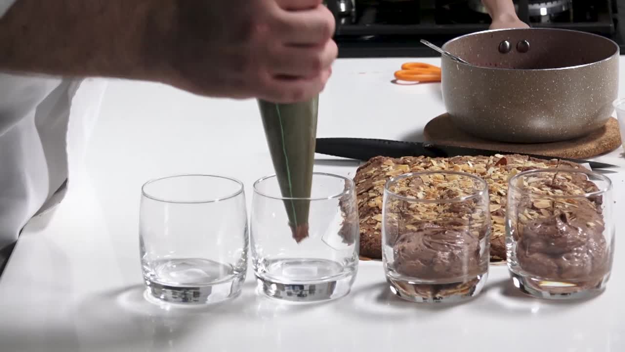 A chef skillfully pipes creamy chocolate mousse into glasses while preparing a dessert on a clean kitchen countertop. The atmosphere is inviting and culinary