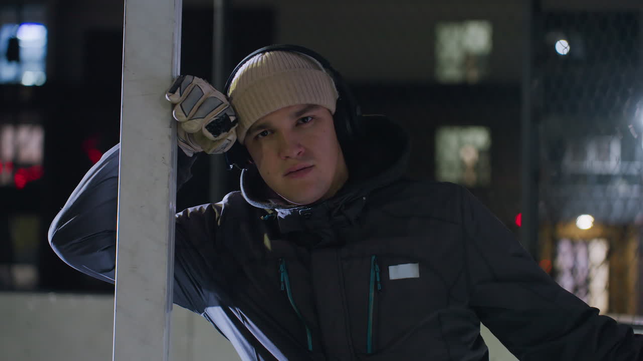 Close-up of man in hoodie resting on pole under bright night lighting, wearing gloves and a beanie, with headphones, chain-link fence in background, and urban lights illuminating the scene