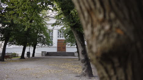 Old-white-building-with-weathered-wooden-doors-sits-in-a-park-of-trees-covering-the-walkway