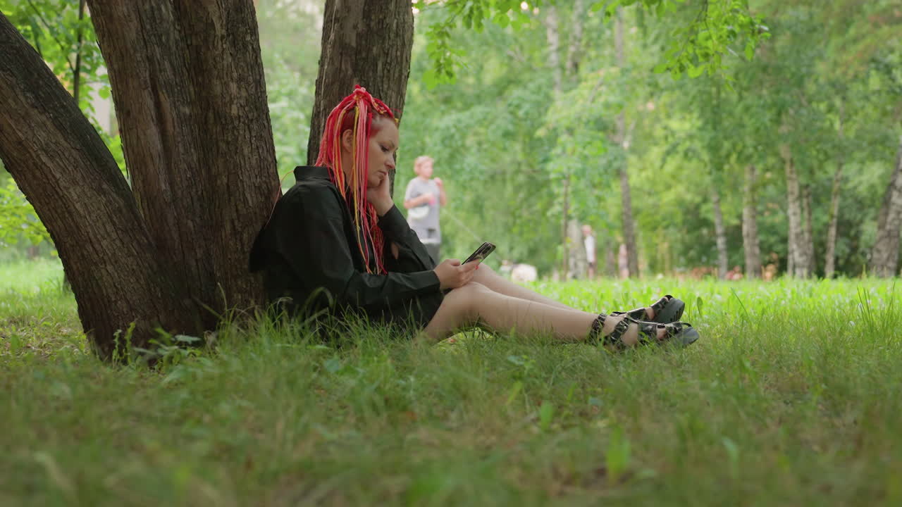 Спокойная женщина отдыхает под деревом, наблюдая за другими; безмятежная женщина с красными дредами смотрит на пробегающего мимо бегуна в парке; кроткая женщина со спокойным взглядом остается сидеть, пока бегун тихо пробегает мимо в парке.