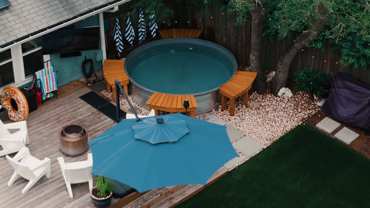 Aerial close drone pull away of a hot tub pool with deck umbrella, white Adirondack chairs, and oak shade. Outdoor living dream home, real estate listings, and home property content