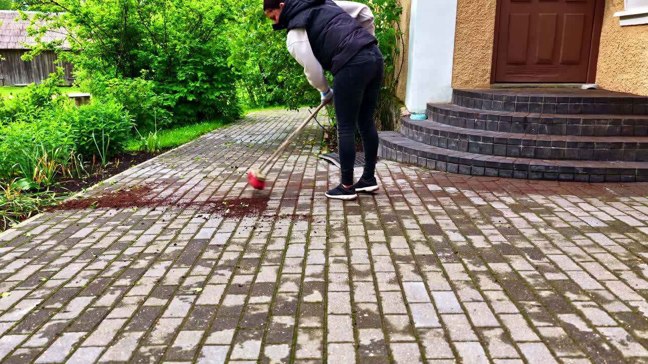 A woman does yard work, using a push broom to sweep up a pile of wet soil from the interlocking paving stones of her house's backyard patio after gardening