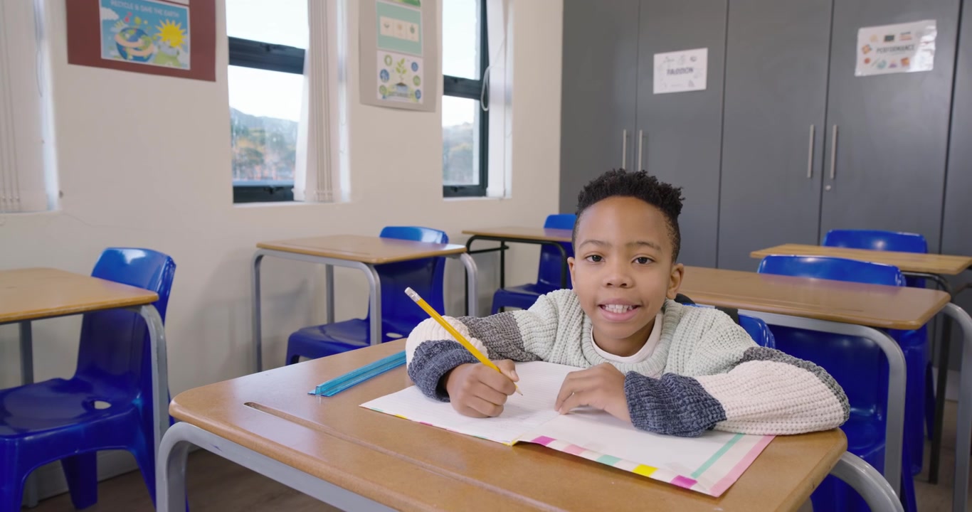 Bambino africano in età scolare che scrive con una matita gialla su un libro al banco in classe, mentre la telecamera si avvicina.