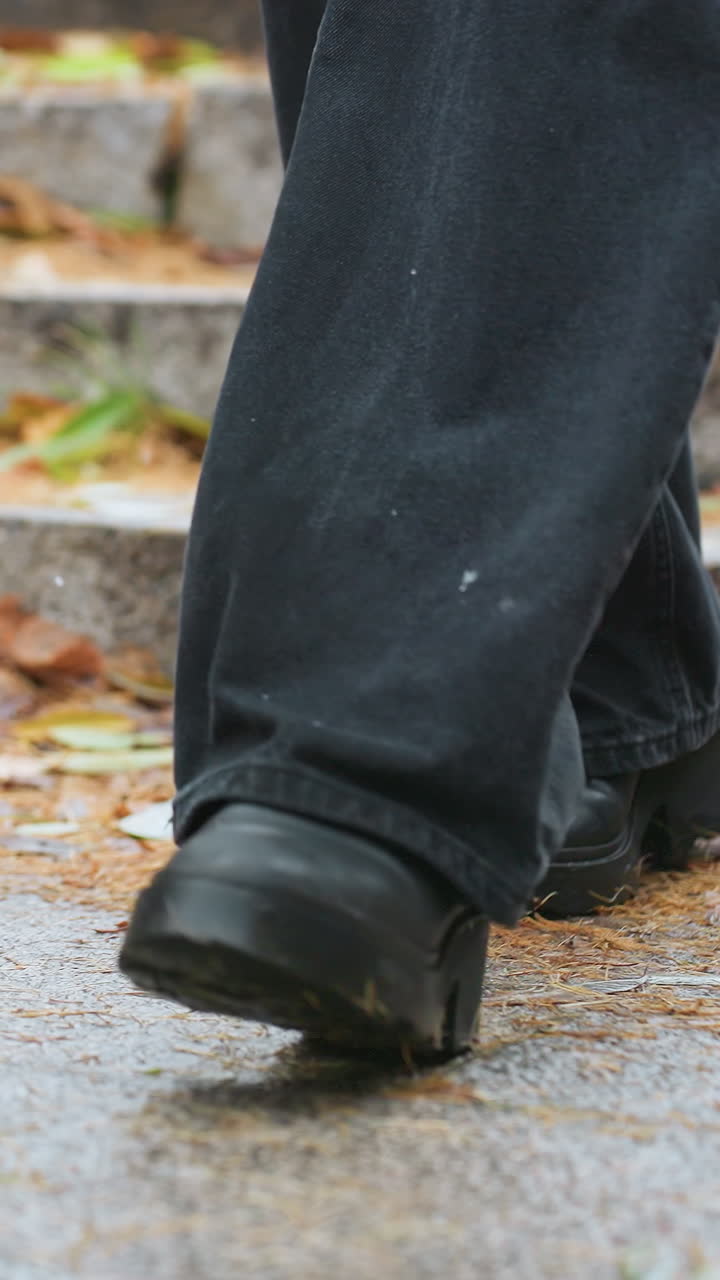 Human legs walking down stone steps during light snowfall, wearing black jeans and black boots, with colorful autumn leaves scattered along the path creating a seasonal vibe