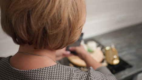 back view of mother with short brown hair raising smartphone to capture image of faintly visible decorated dessert setup on kitchen table with scattered shells and gold utensils in modern interior