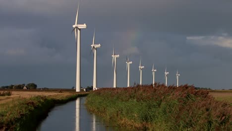 een rij draaiende windturbines met een afwateringssloot op de voorgrond en een regenboog en stormachtige lucht