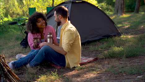 happy couple have a hot coffee with a thermos and a metal mug camping in the forest. caucasian man and young black female outdoors.