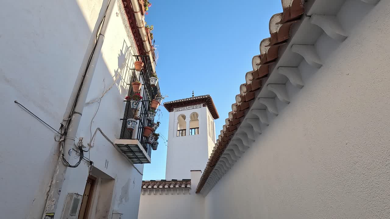 Mezquita Mayor de Granada'nın zarif kulesi, geleneksel Endülüs mimarisi ve canlı saksı bitkileriyle çevrili, berrak mavi bir gökyüzüne karşı yükseliyor. Granada'nın İslami güzelliği.