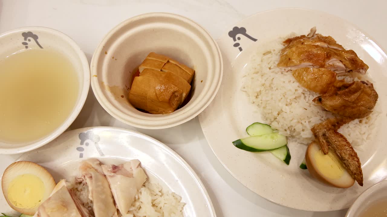 Overhead view of Hainanese chicken rice and roasted chicken rice meals being served, with soup, braised egg, tofu, and garnishes under bright indoor lighting