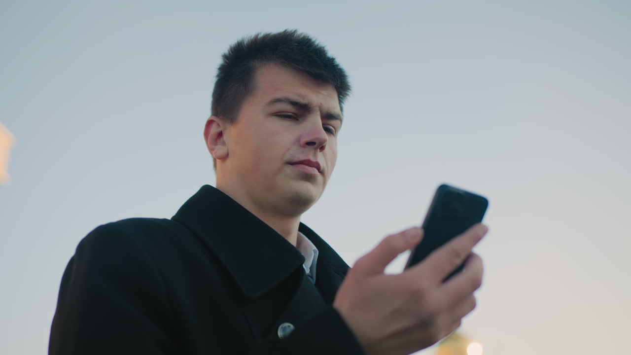 검은 코트와  셔츠를 입은 전문적인 남자가 은 도시 스카이라인을 배경으로 밖에서 드폰을 쳐다보고 집중과 결단력을 반영하고 있습니다.