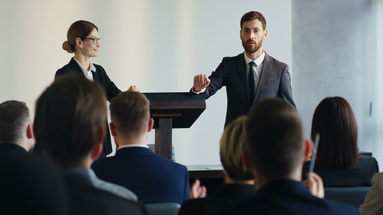 кавказский мужчина выступает с речью на конференции и отвечает на вопросы слушателей, его коллега-женщина стоит рядом с ним на трибуне