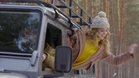 a young and pretty woman leaning out of a cars window enjoying the surrounding and fresh air on her face whilst her boyfriend drives