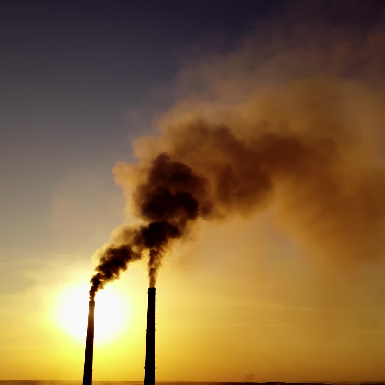 Two industrial pipes with dark smoke at sunset. Massive dense of toxic emissions from industrial factory filling the sky in the evening. Polluting the atmosphere.