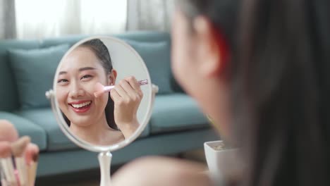 reflection in a mirror beautiful young asian woman rouging her cheeks and smiling while applying makeup at home