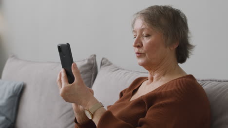 Senior-Woman-Using-A-Smartphone-While-She-Is-Sitting-On-Sofa-In-Living-Room-At-Home