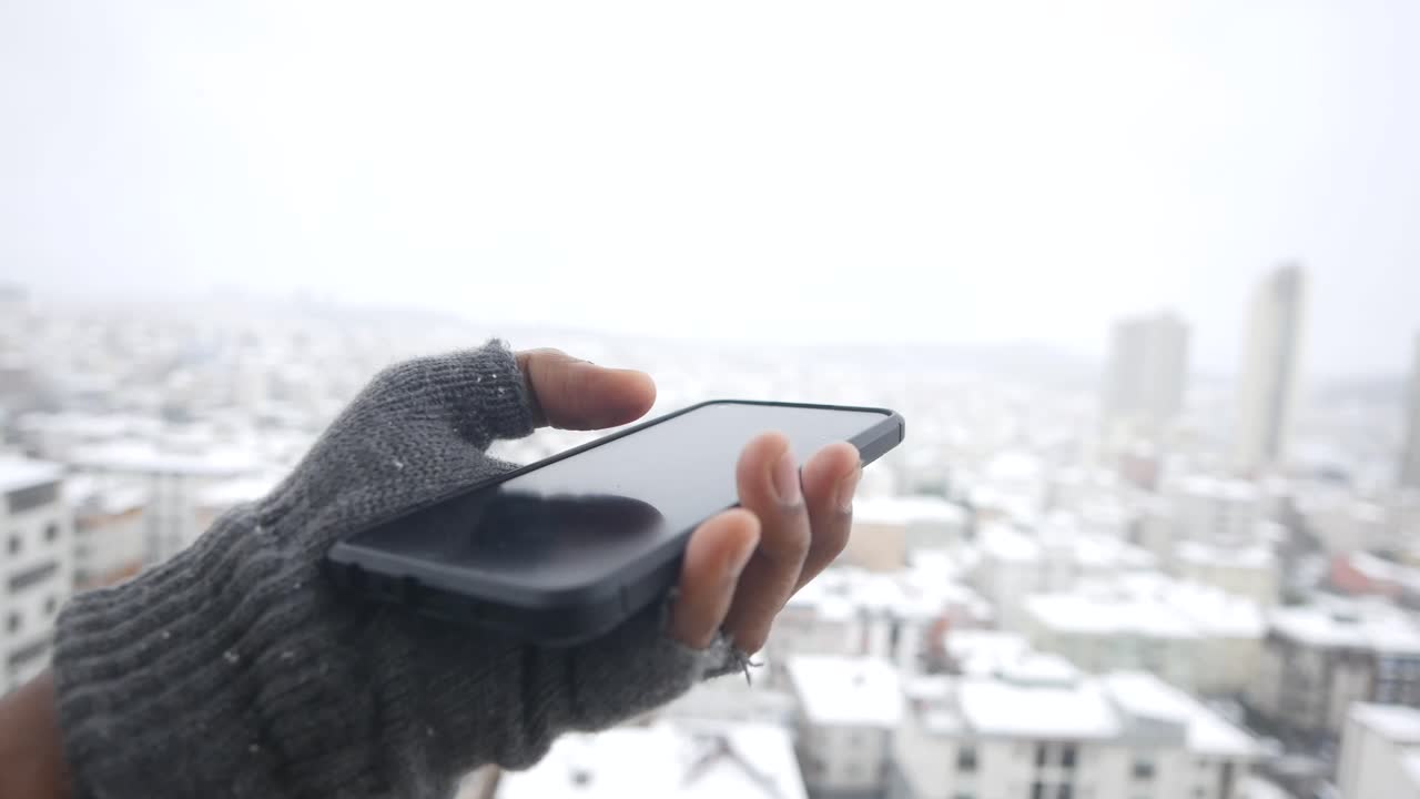 Hand in Glove Holding Smartphone with Snowy City Background