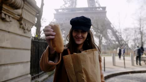 beautiful happy european woman walking near eiffel tower, giving a piece of french baguette to camera man slow motion.