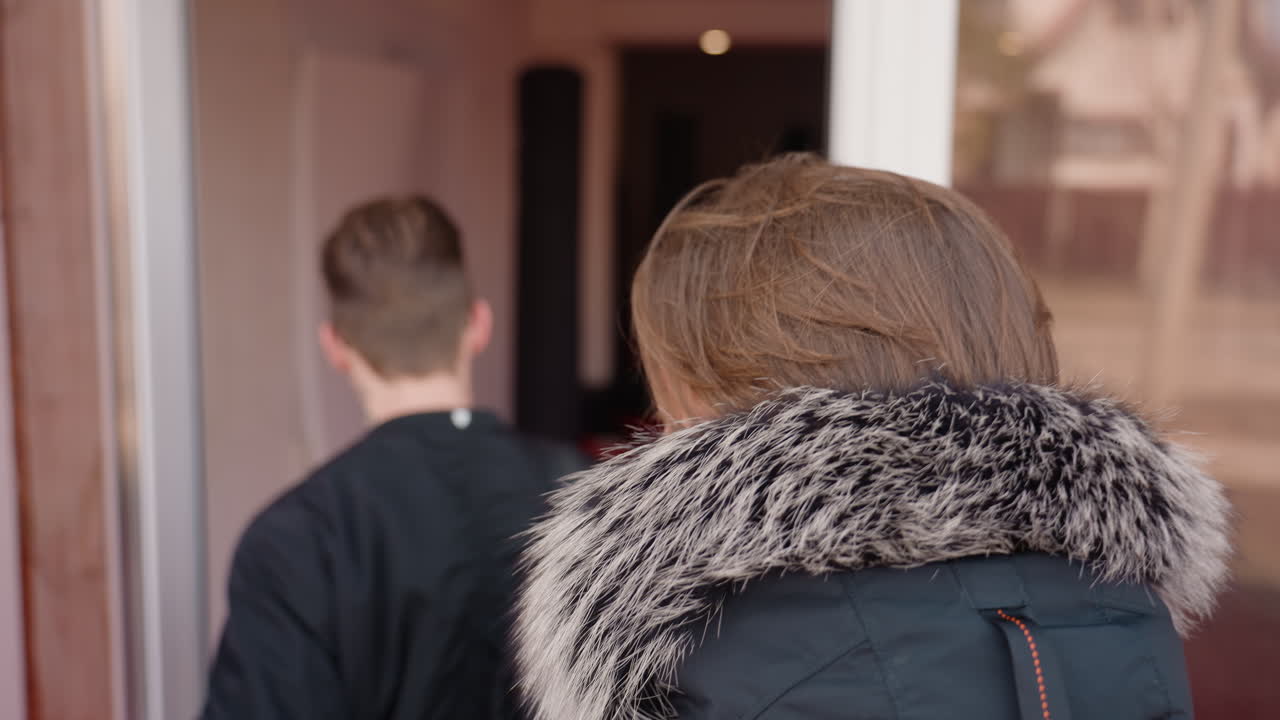 Two guys in jackets walking into glass entrance of wooden building, one carrying black backpack, other with fur hood coat, reflection visible on glass door, moment of pause before entering urban environment