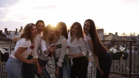 emotional bridesmaid and attractive girlfriends talking selfie with mobile phone and smiling. caucasian girls in white shitrs posing and holding flowers in hands. outdoors. front view