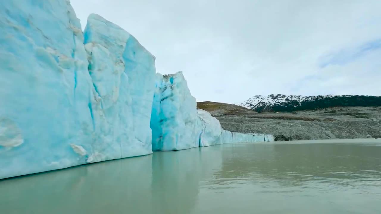 FPV aerial drone gliding and flying close to icy blue and tall steep glaciers that are in Greenland or Iceland water during a cloudy day. The water is brown and landscape with trees in the background.