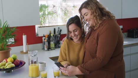 Happy-caucasian-lesbian-couple-embracing-and-using-smartphone-in-kitchen