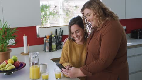 feliz pareja de lesbianas caucásicas abrazándose y usando un teléfono inteligente en la cocina