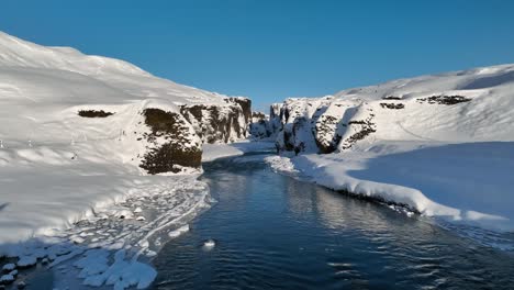 Aerial-view-over-water-from-a-glacier-river-flowing-through-a-canyon-covered-in-snow,-on-a-sunny-day