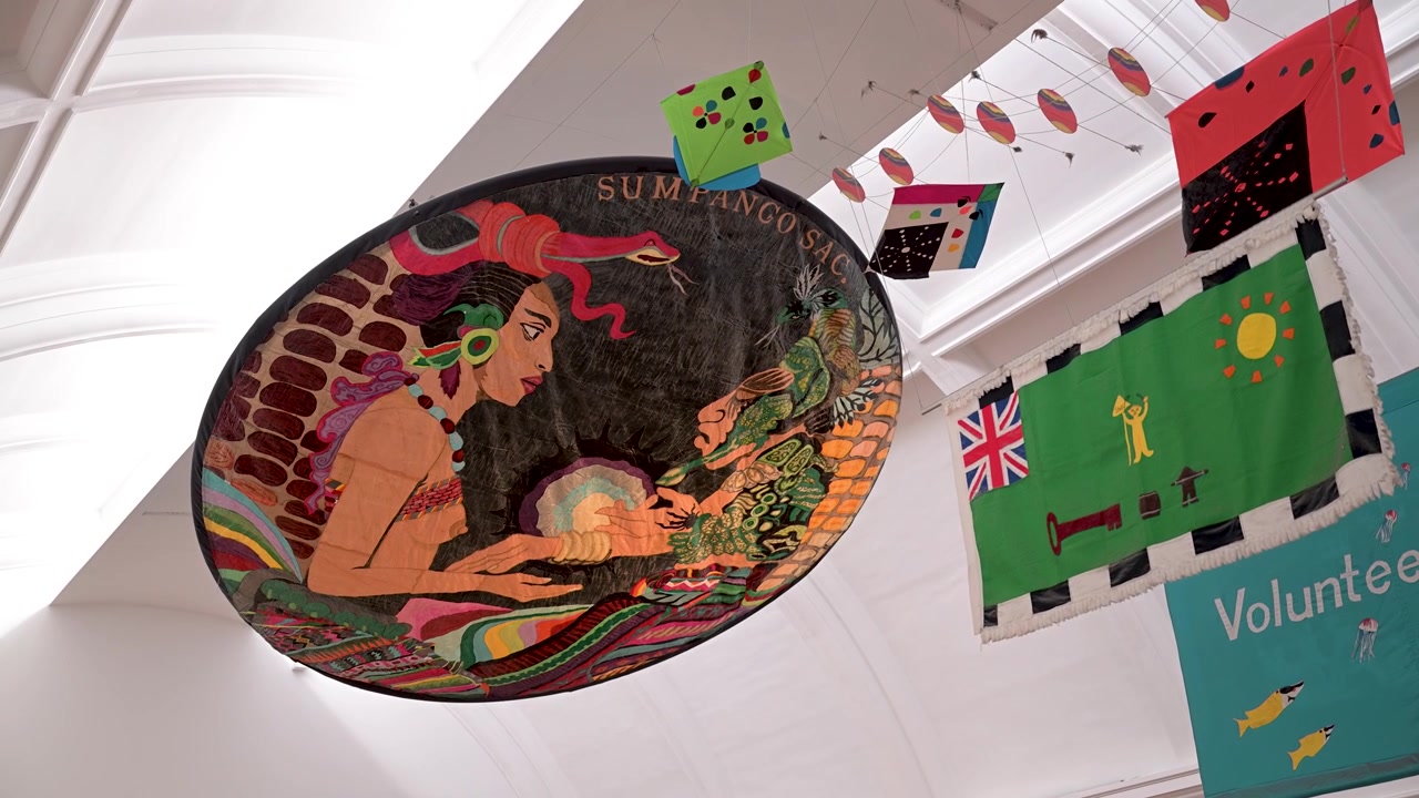 Large circular artwork and bright fabric banners hang from the high ceiling of a museum hall in a slow upward pan.