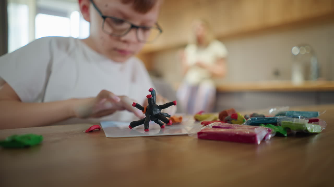 Boy with glasses focused on sculpting colorful clay at wooden table with black clay figure in foreground, packets of modeling clay scattered, mother blurred in background, symbolizing creativity, concentration