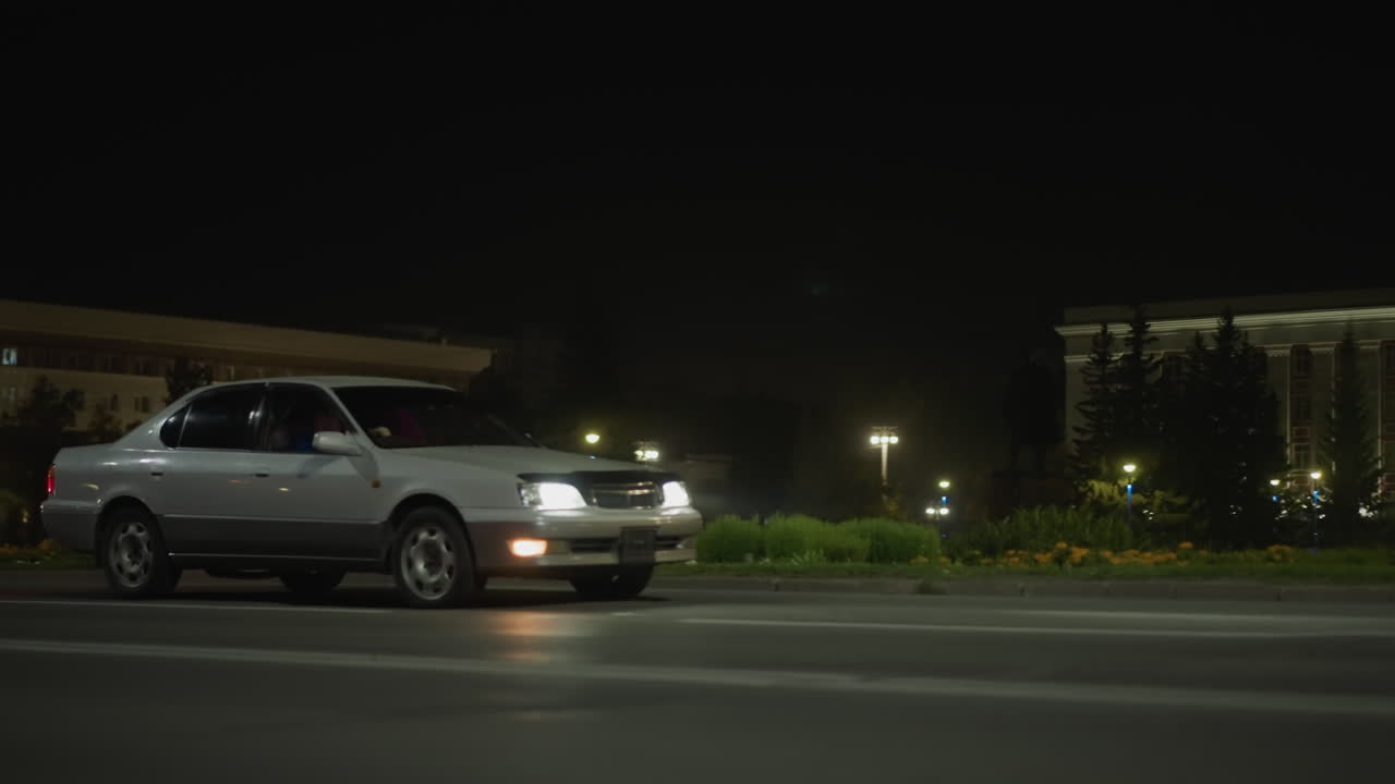 다가오는 헤드라이트와 세단이 있는 밤의 교차로, 가로등과 빌보드 불빛이 있는 텅 빈 시내 대로, 젖은 아스팔트의 반사와 희미한 안개, 전경의 길가 기둥, 느린 영화 같은 장면