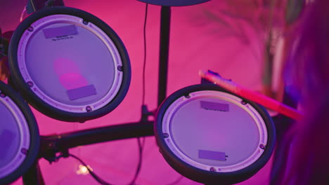 partial view of person using drumsticks to strike electronic drum pads illuminated by vivid neon lighting, soft focus creating artistic mood with cables and setup visible in colorful background