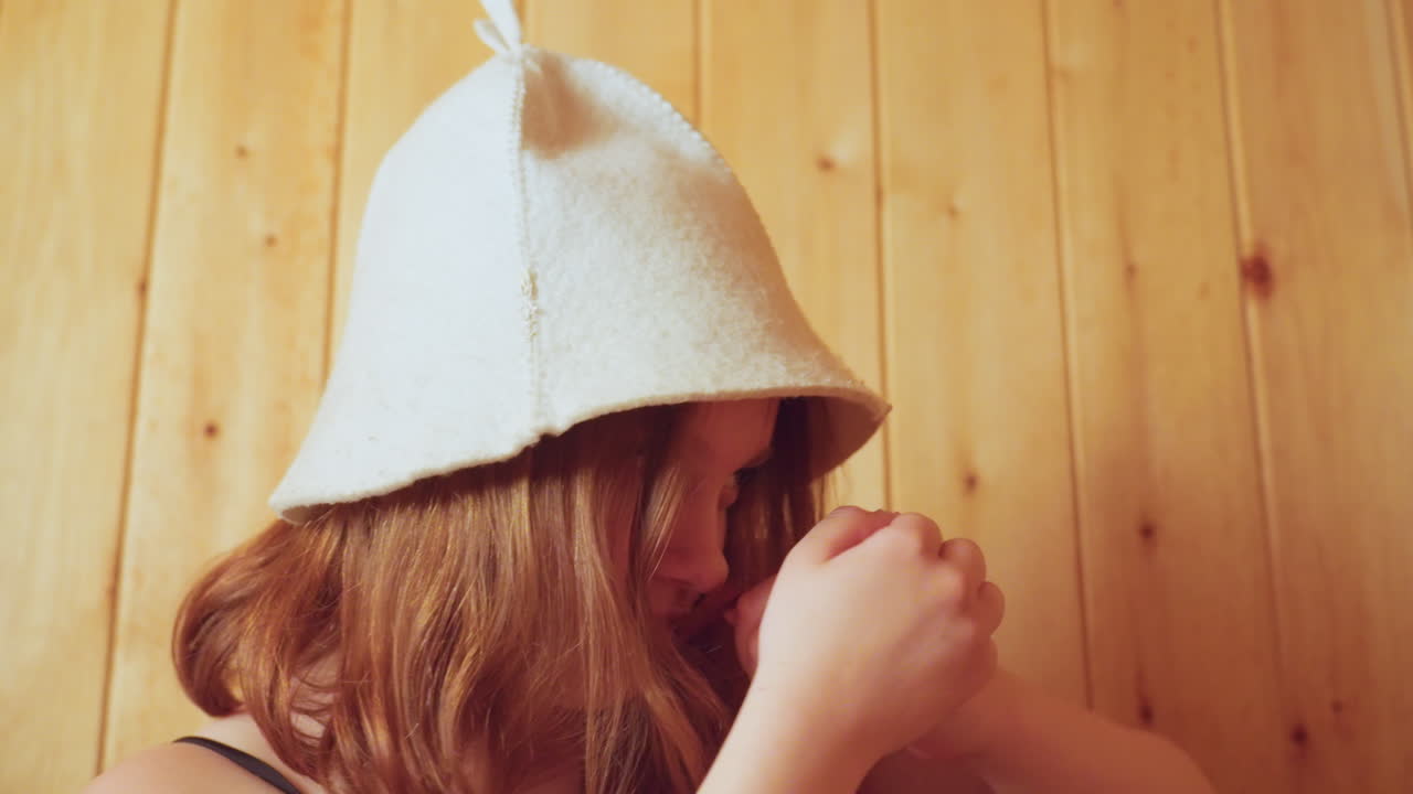 Young lady wearing soft white sauna cap lifts both hands in playful gesture while standing against wooden wall, smiling subtly with relaxed confident energy and having fun indoors