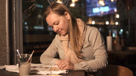 una chica rubia está pintando en una camiseta blanca con un pincel, sentada en un café por la noche